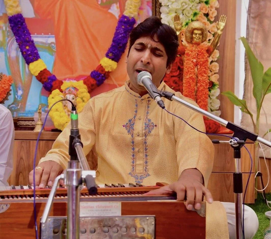 Dr. Koustubh Pare singing with harmonium before a garlanded portrait of Sri Sathya Sai Baba